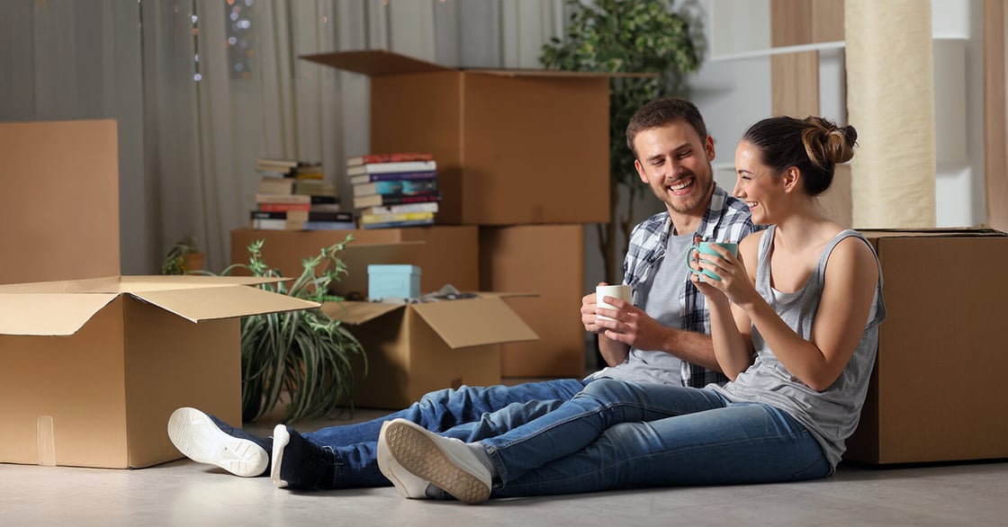 A couple sitting in their new home surrounded by moving boxes, drinking coffee.